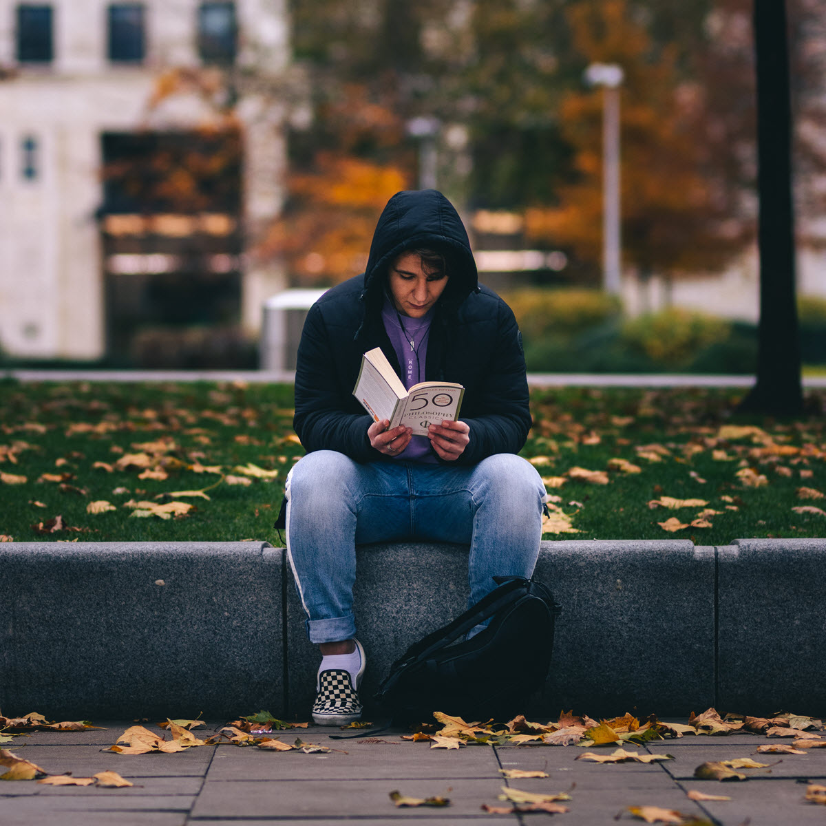 Person reads book outdoors, surrounded by fallen autumn leaves. Photo credit Jordan Brierley at Unsplash